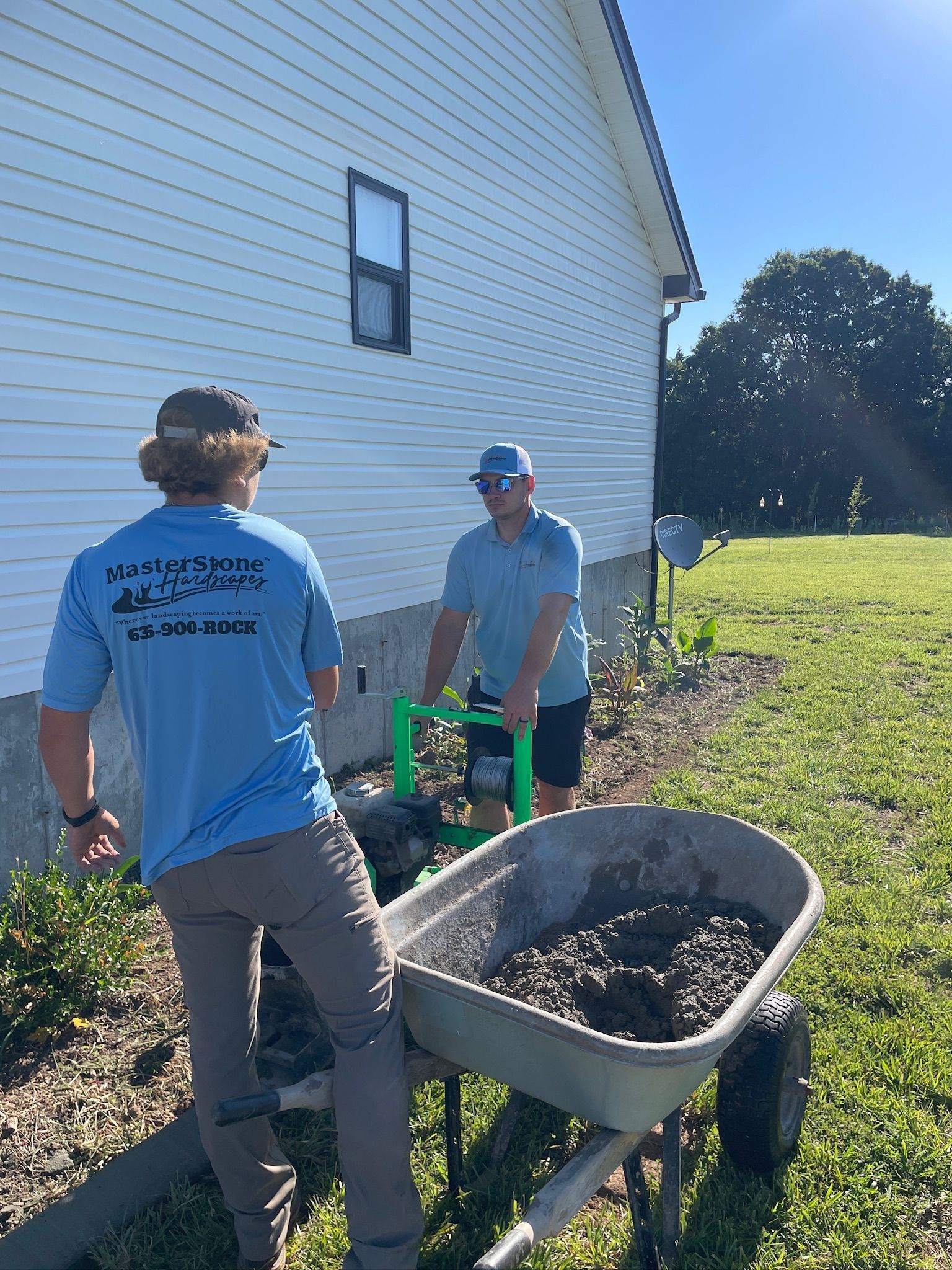 Two people moving a wheelbarrow of mulch beside a white house on a sunny day