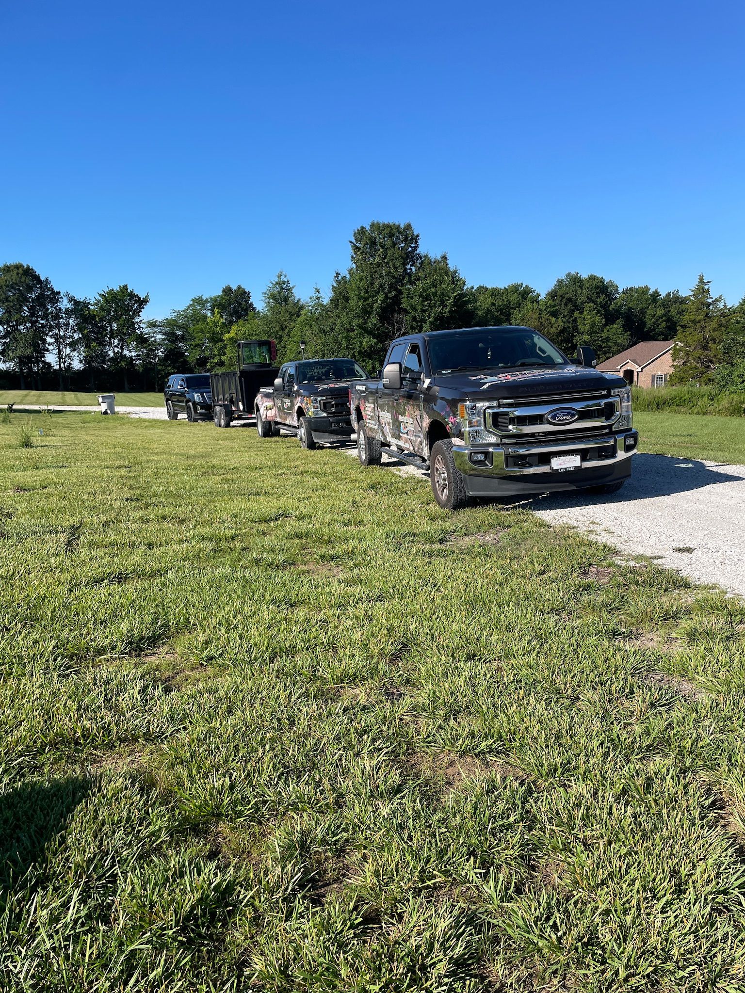 Line of black pickup trucks parked on a gravel drive beside a grassy field under a clear blue sky