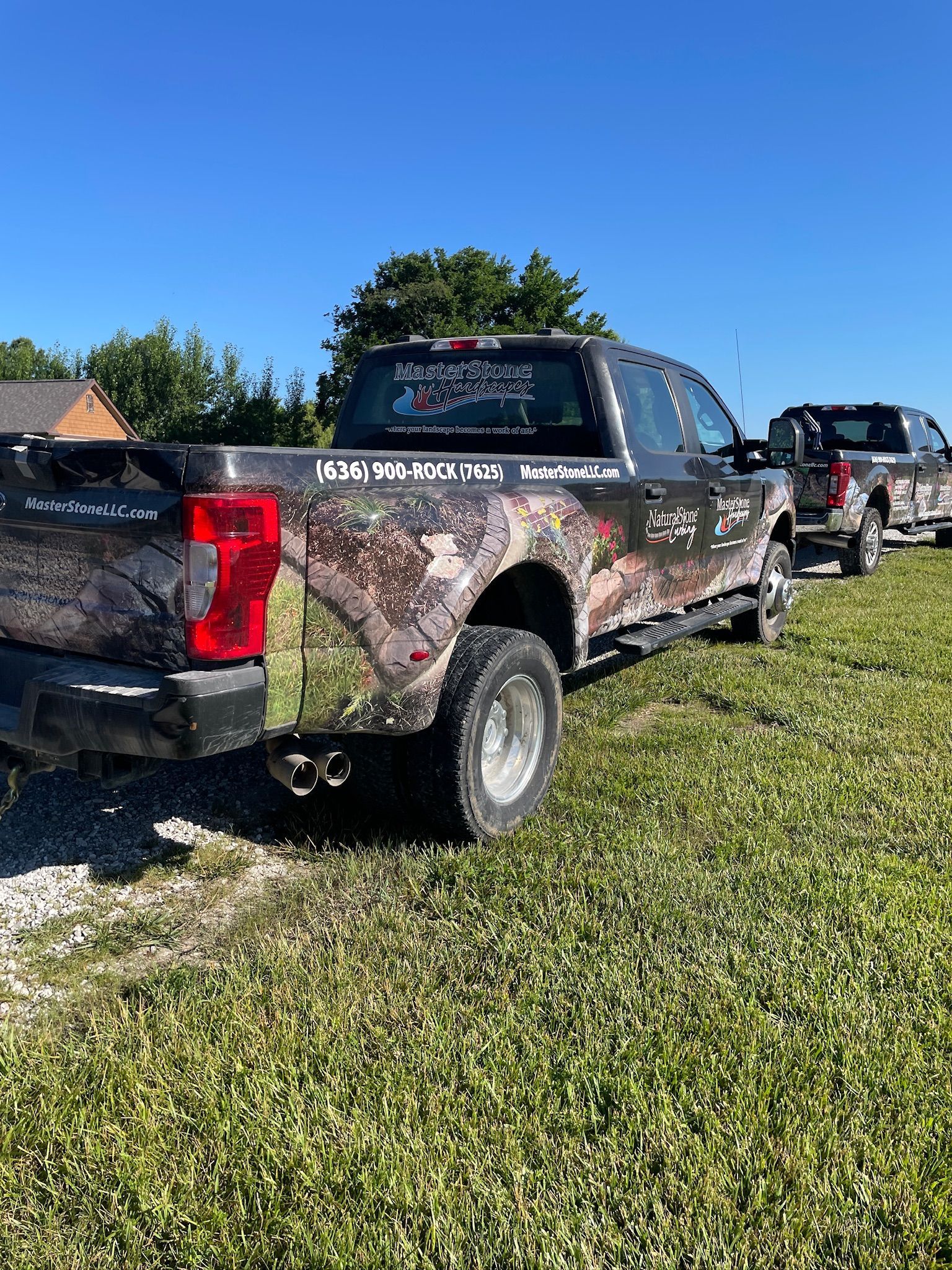 Camouflage pickup truck parked on grass under a clear blue sky, with another truck behind it