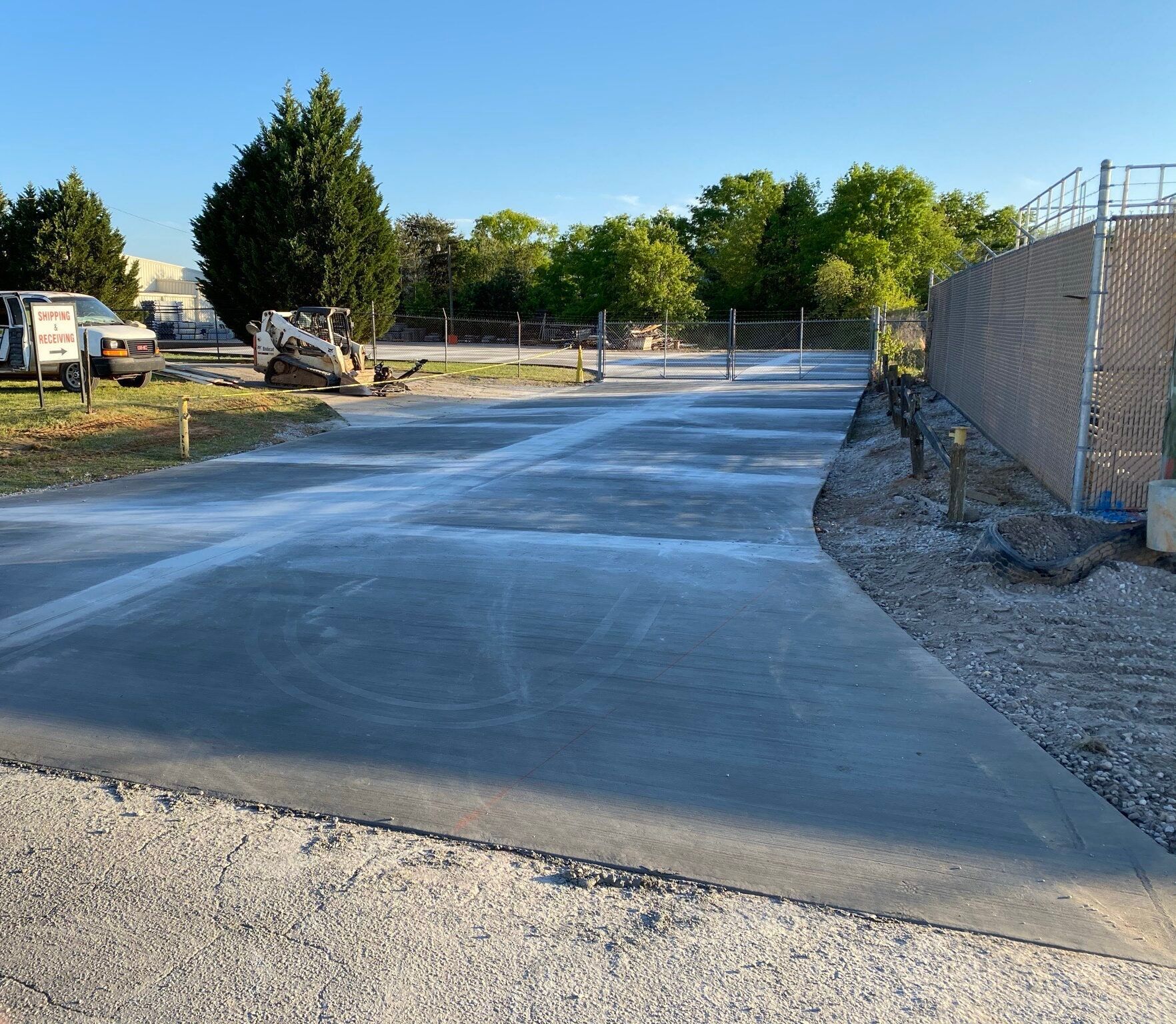 Newly poured concrete driveway with a gravel edge, near a chain-link fence and trees.