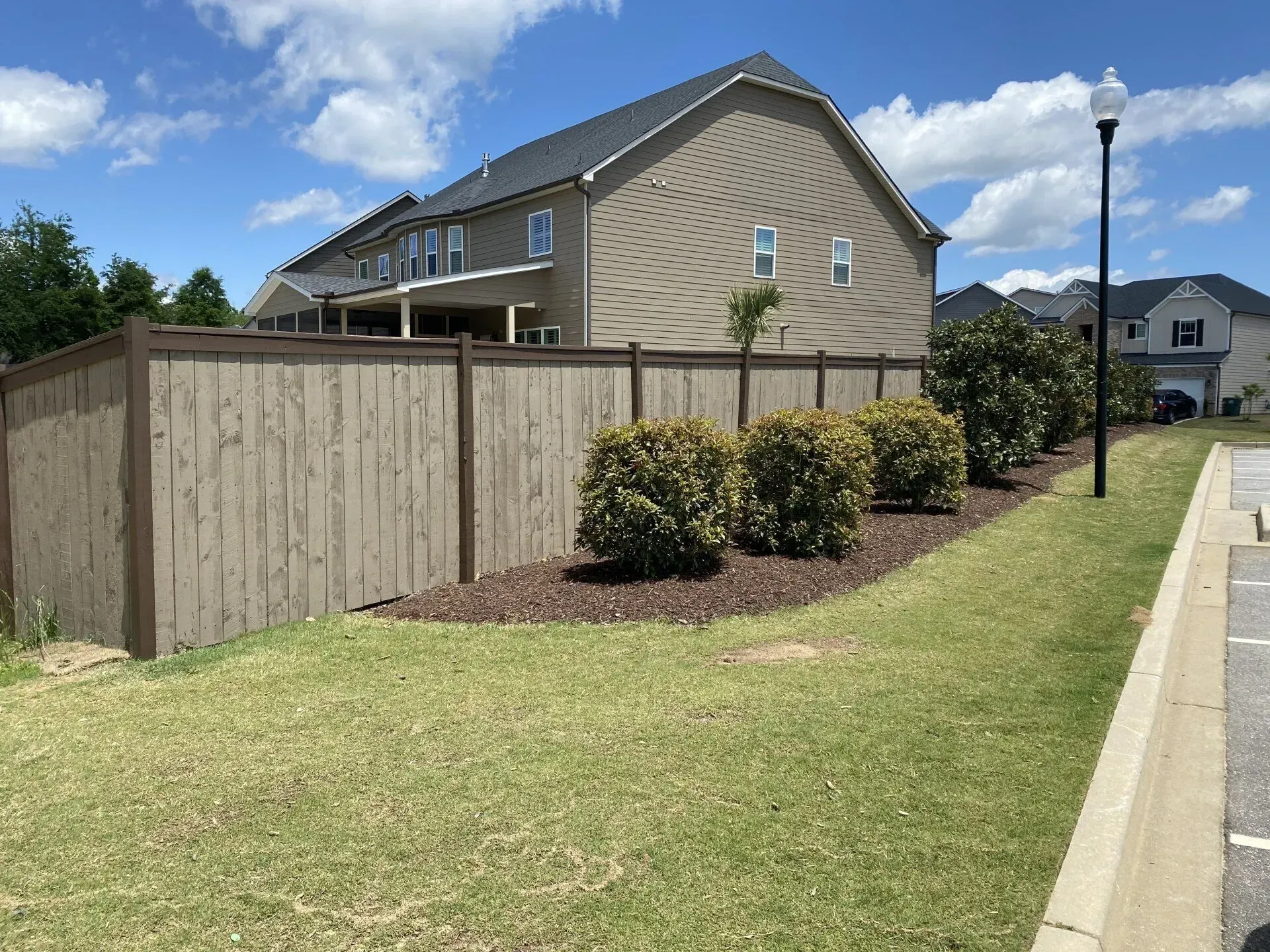 Tan wooden fence, manicured lawn, shrubs, beige house, blue sky with clouds.
