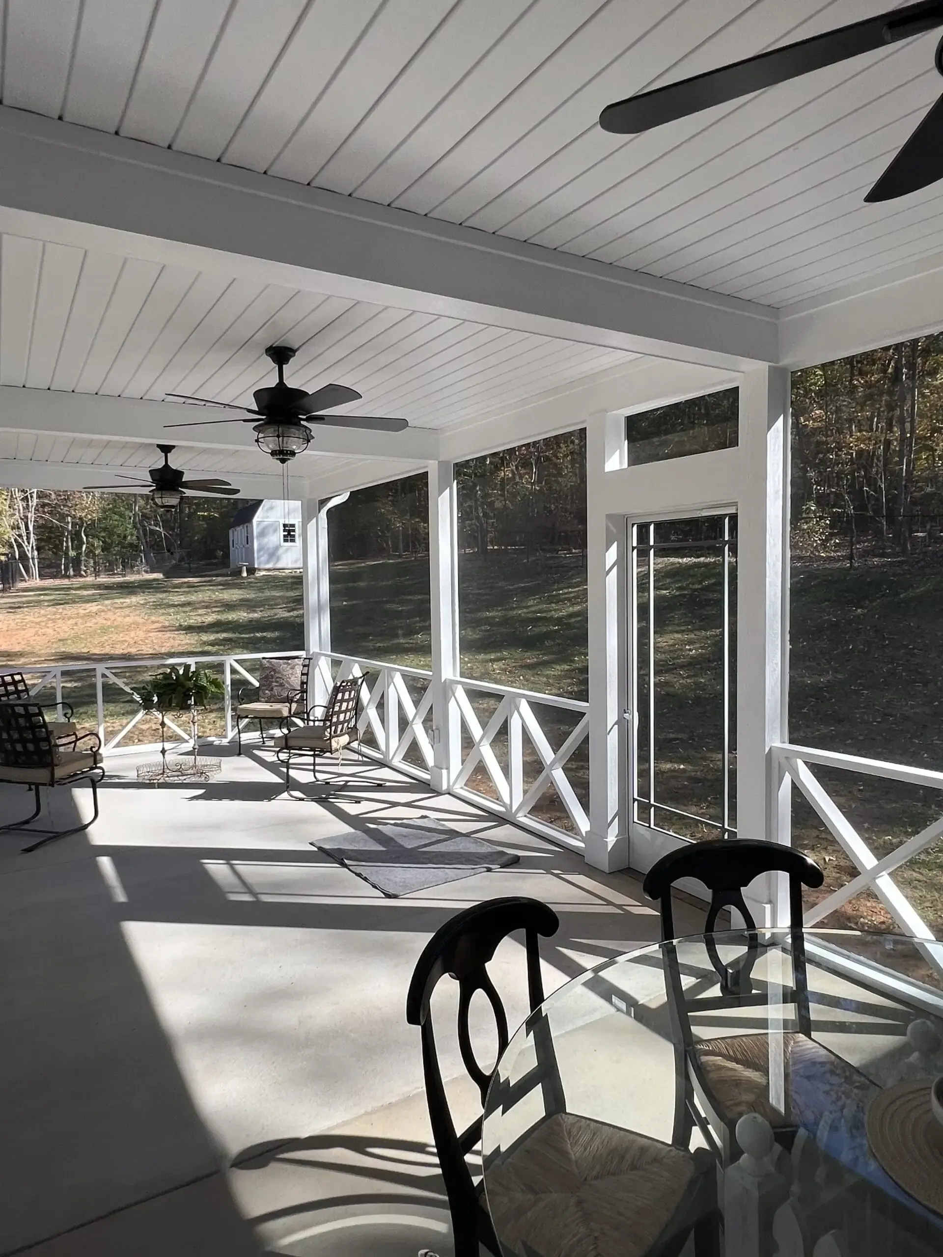 Screened-in porch with white ceiling, ceiling fans, and X-shaped railing. Sunlight streams in, illuminating the concrete floor.