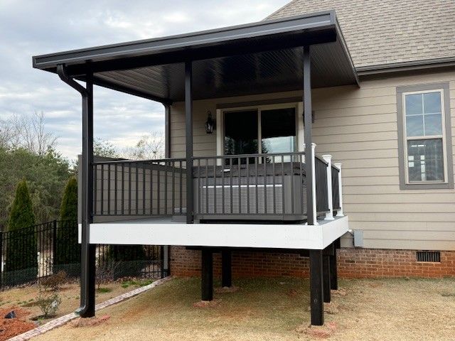 Dark-framed, covered deck attached to a house with a white base and a gray railing, on a lawn.