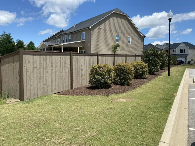 Tan fence along a grassy area, shrubs and a two-story house with blue sky in the background.