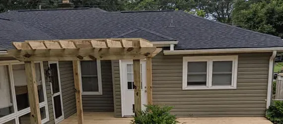 A house with a dark roof and tan siding. There is a wooden pergola in the foreground.