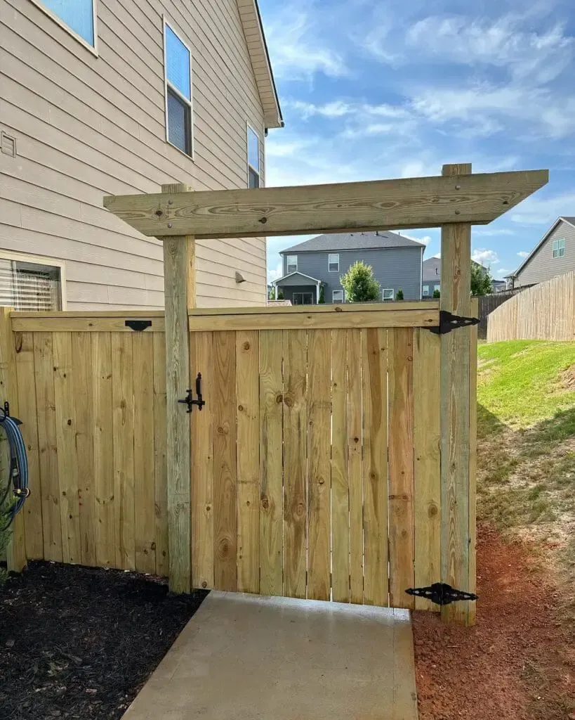 Wooden gate with a pergola, leading to a concrete path in a yard.