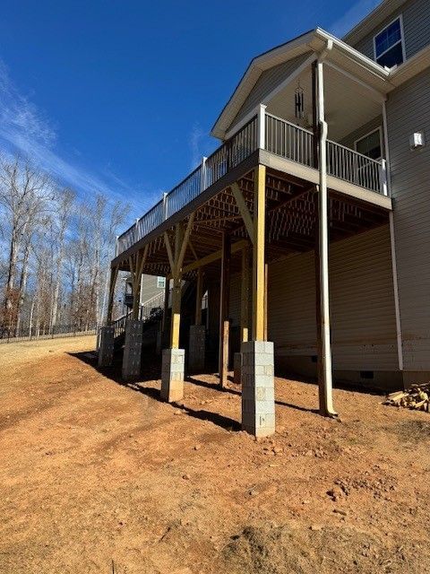Deck attached to a two-story house, supported by pillars, on a dirt lot under a blue sky.