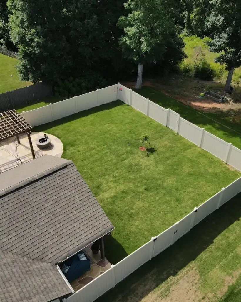 Aerial view of a fenced backyard with green grass, a patio, and trees in the background.
