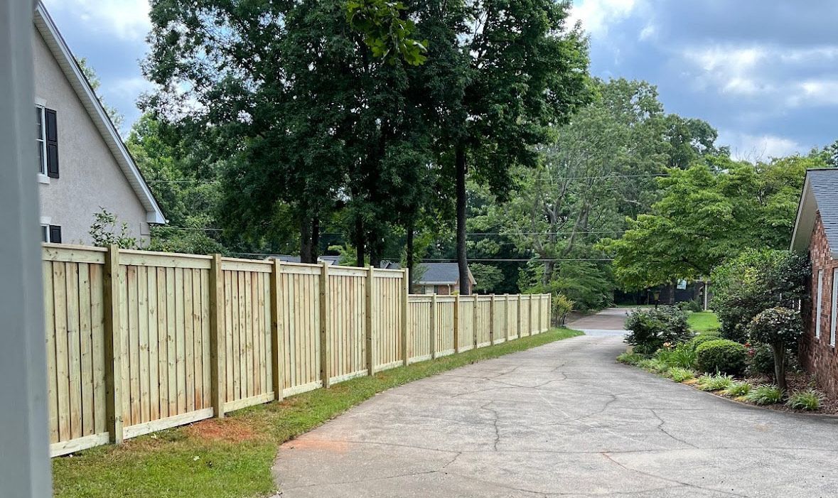 Wooden fence alongside a driveway, trees in background, cloudy sky.