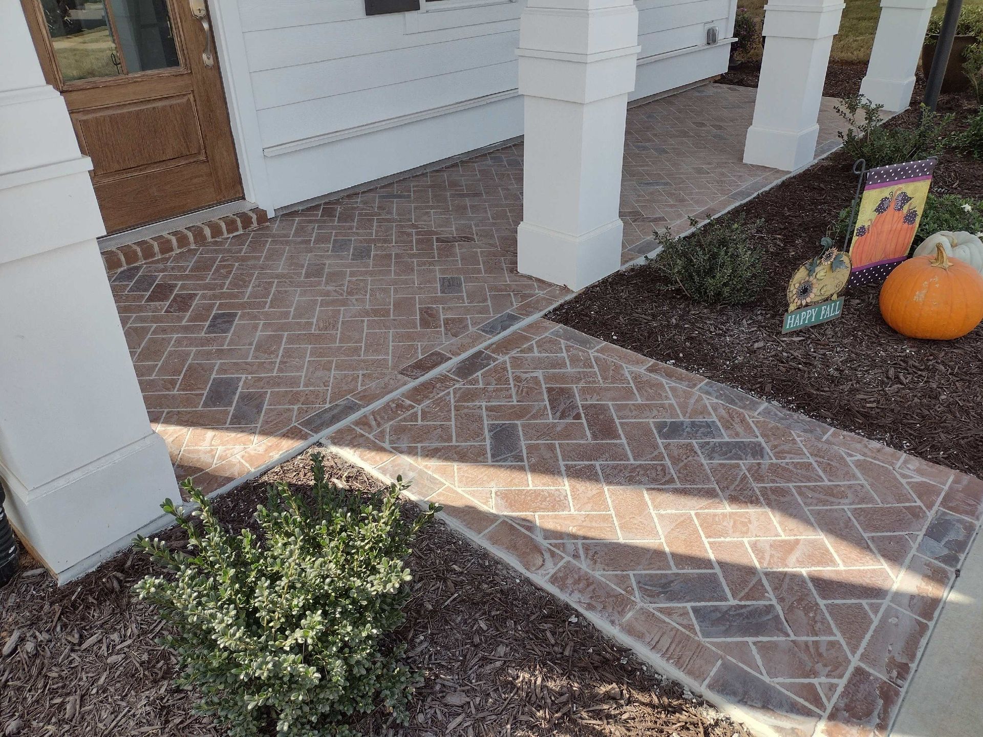 Brick-paved walkway and porch of a house, with white columns, a door, and a Halloween decoration.