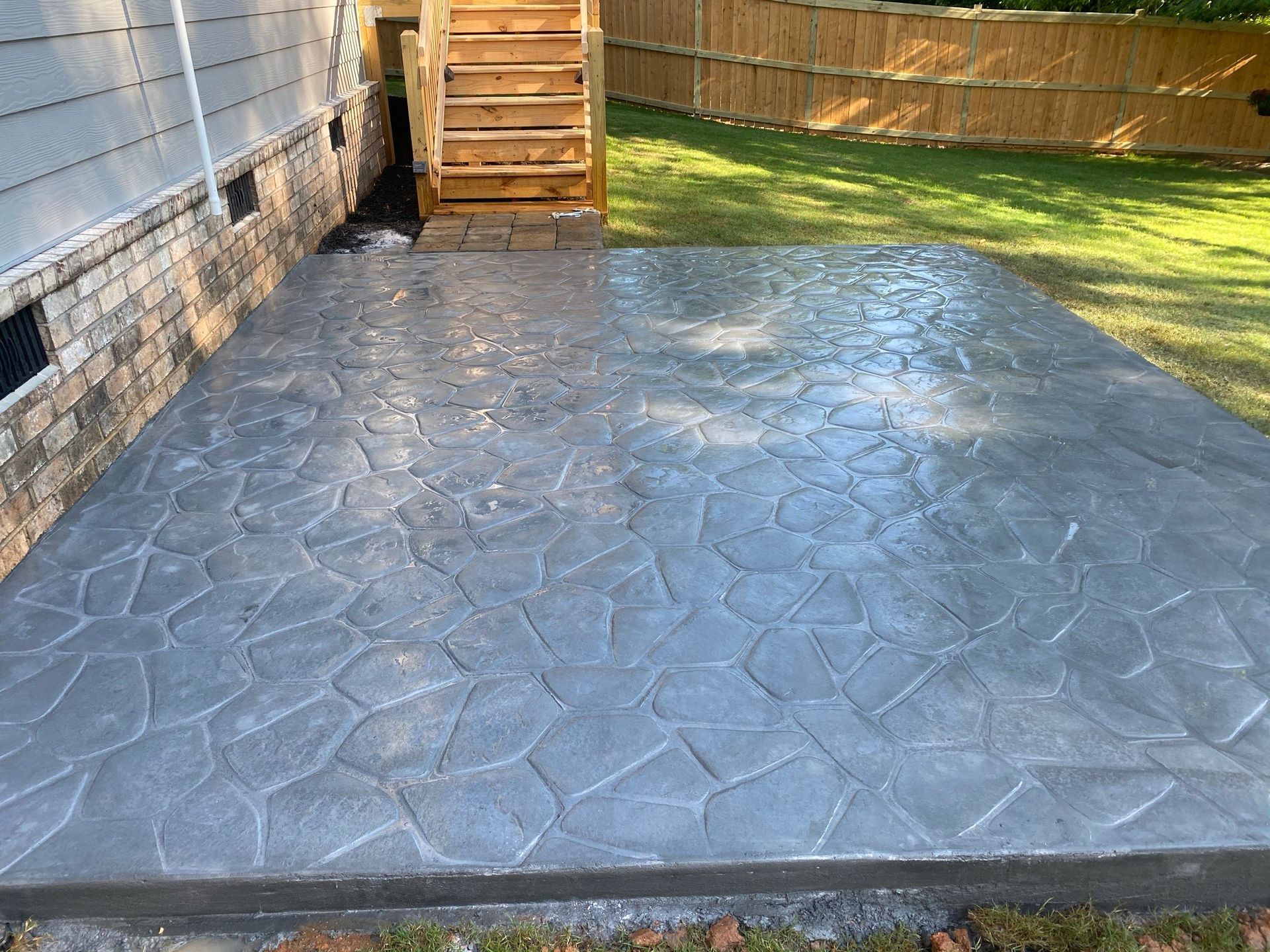 Gray stone-patterned concrete patio next to a house, leading to wooden steps and a fenced lawn.