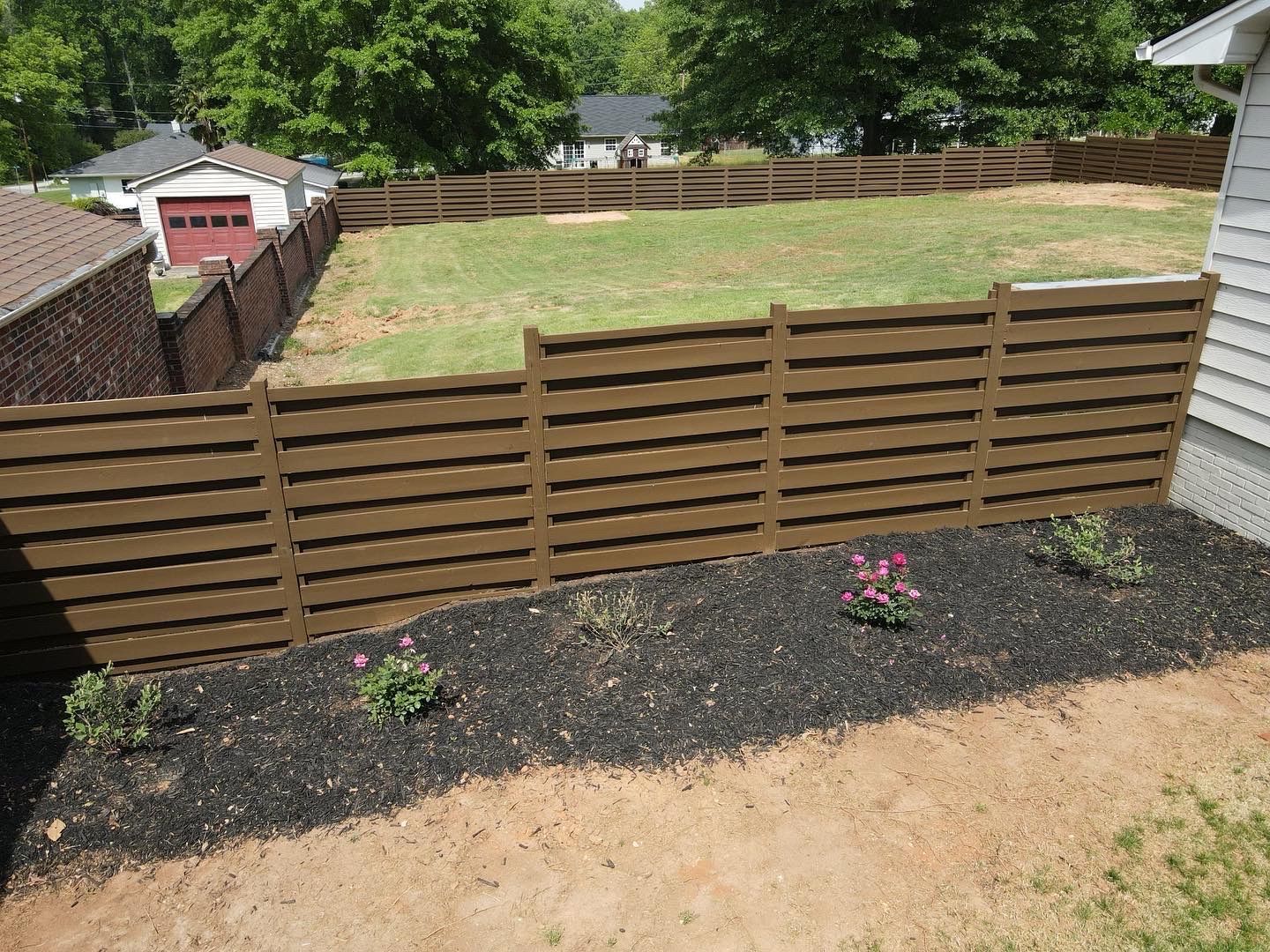 Brown horizontal slat fence encloses a backyard with grass and flowers.