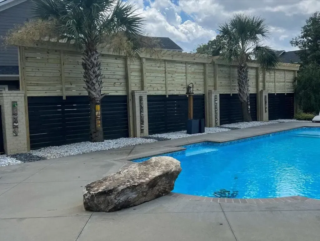 Poolside with black and tan horizontal fence, palm trees, blue water, and large rock.