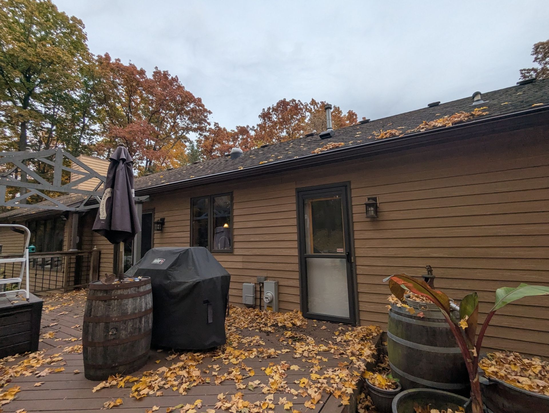 Backyard deck with fallen leaves, barrel planters, grill, and brown siding. Autumn foliage in background.