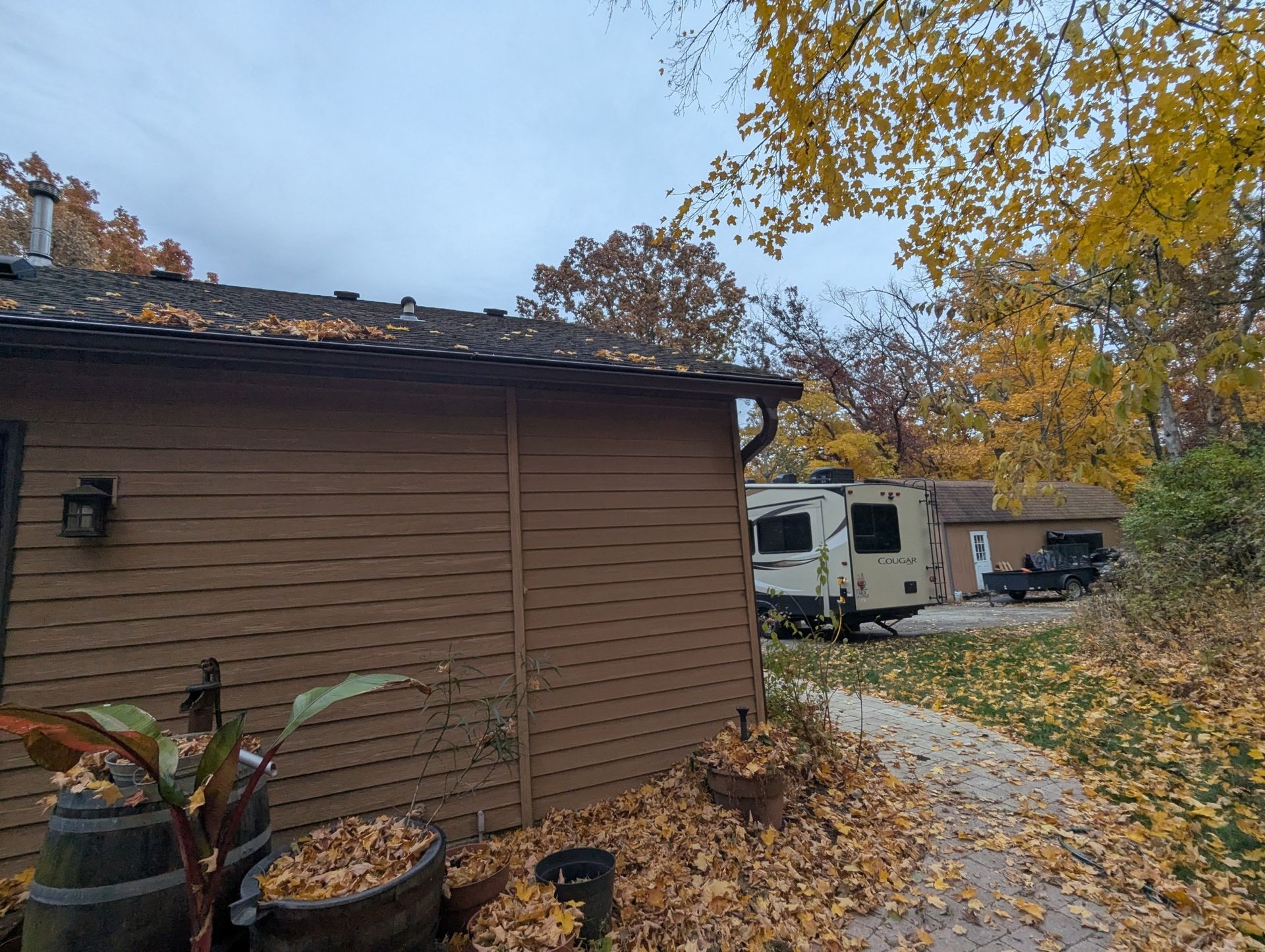 Brown building with a gutter and autumn leaves, RV parked on a driveway with trees. Overcast sky.