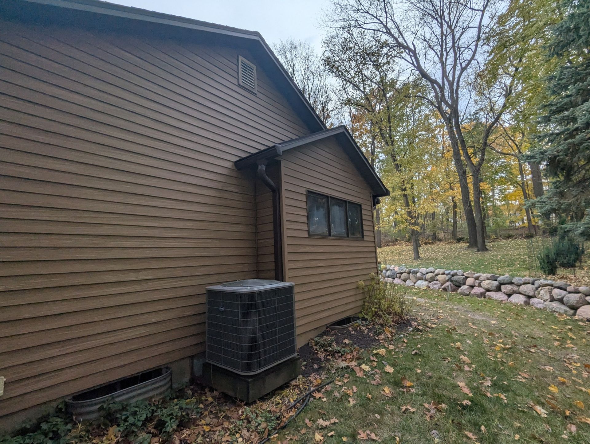 Brown-sided house with a small addition. Air conditioning unit sits next to the addition, with trees and a stone wall in the background.