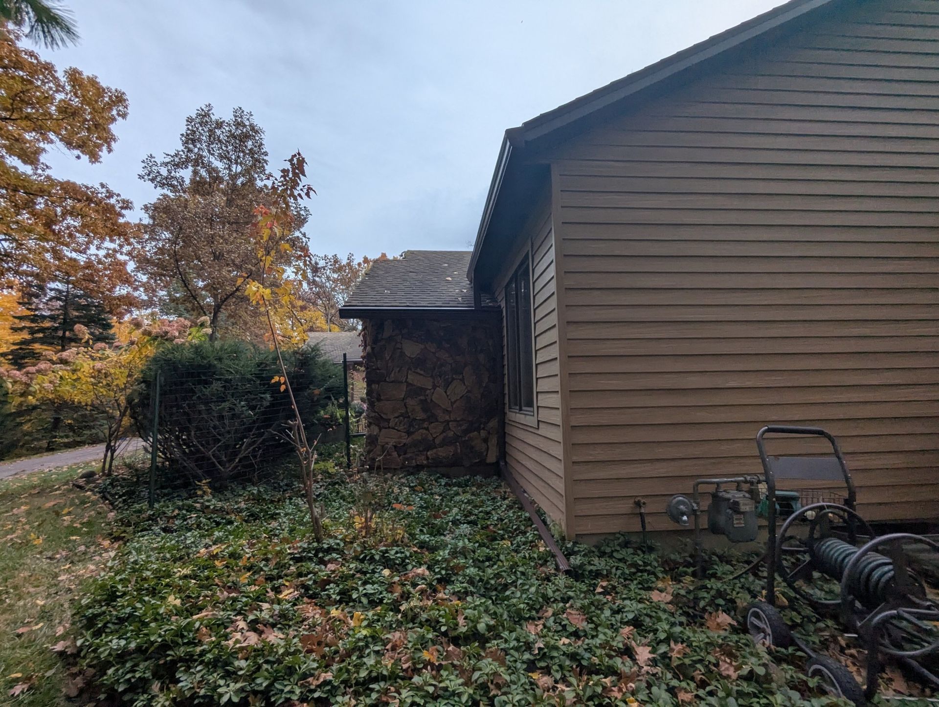 Side of a brown house with bushes, fallen leaves, and a lawnmower. Autumn foliage in the background.