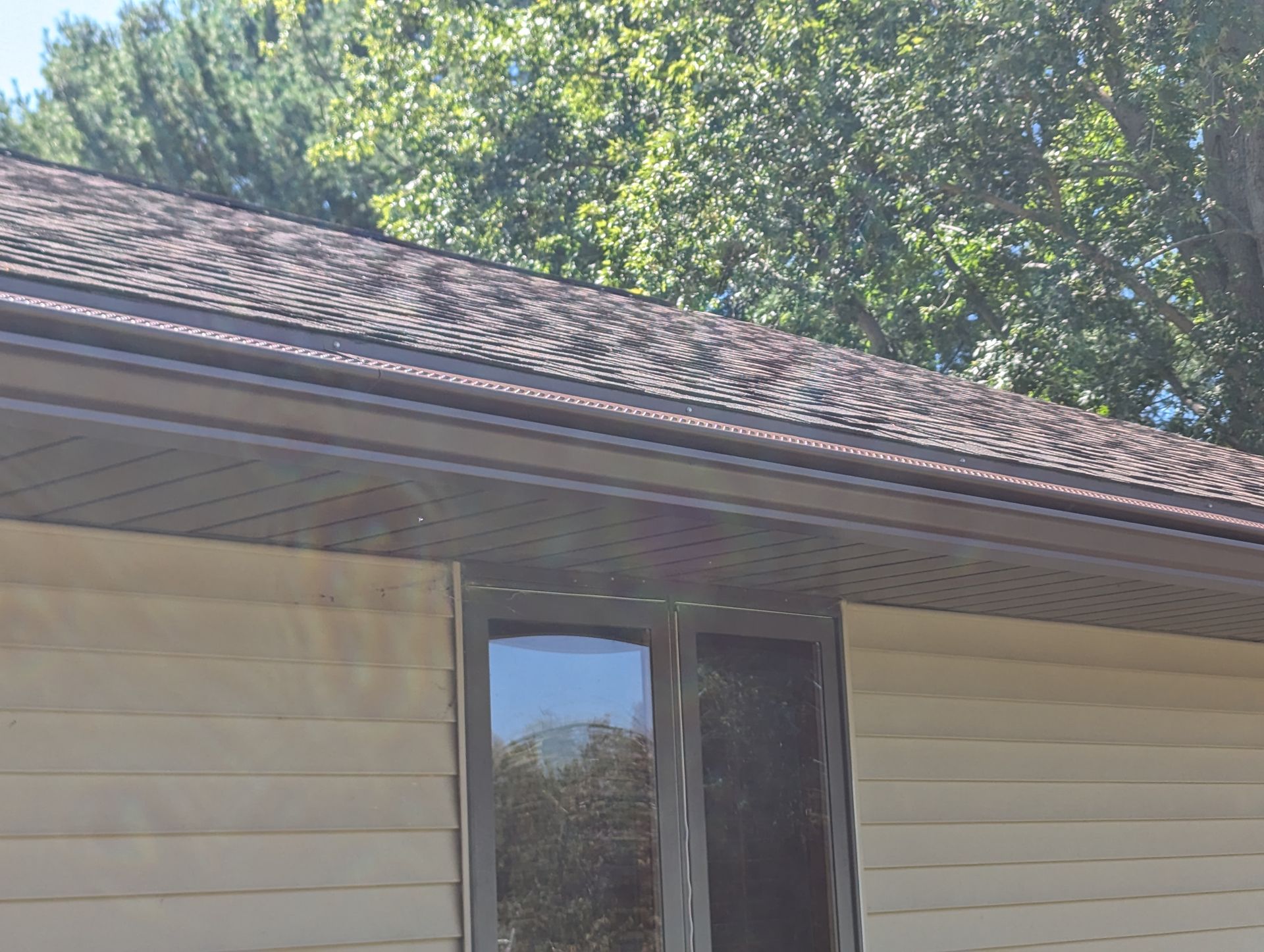 Brown roof, gutters, and siding on a house with a window; trees in the background.