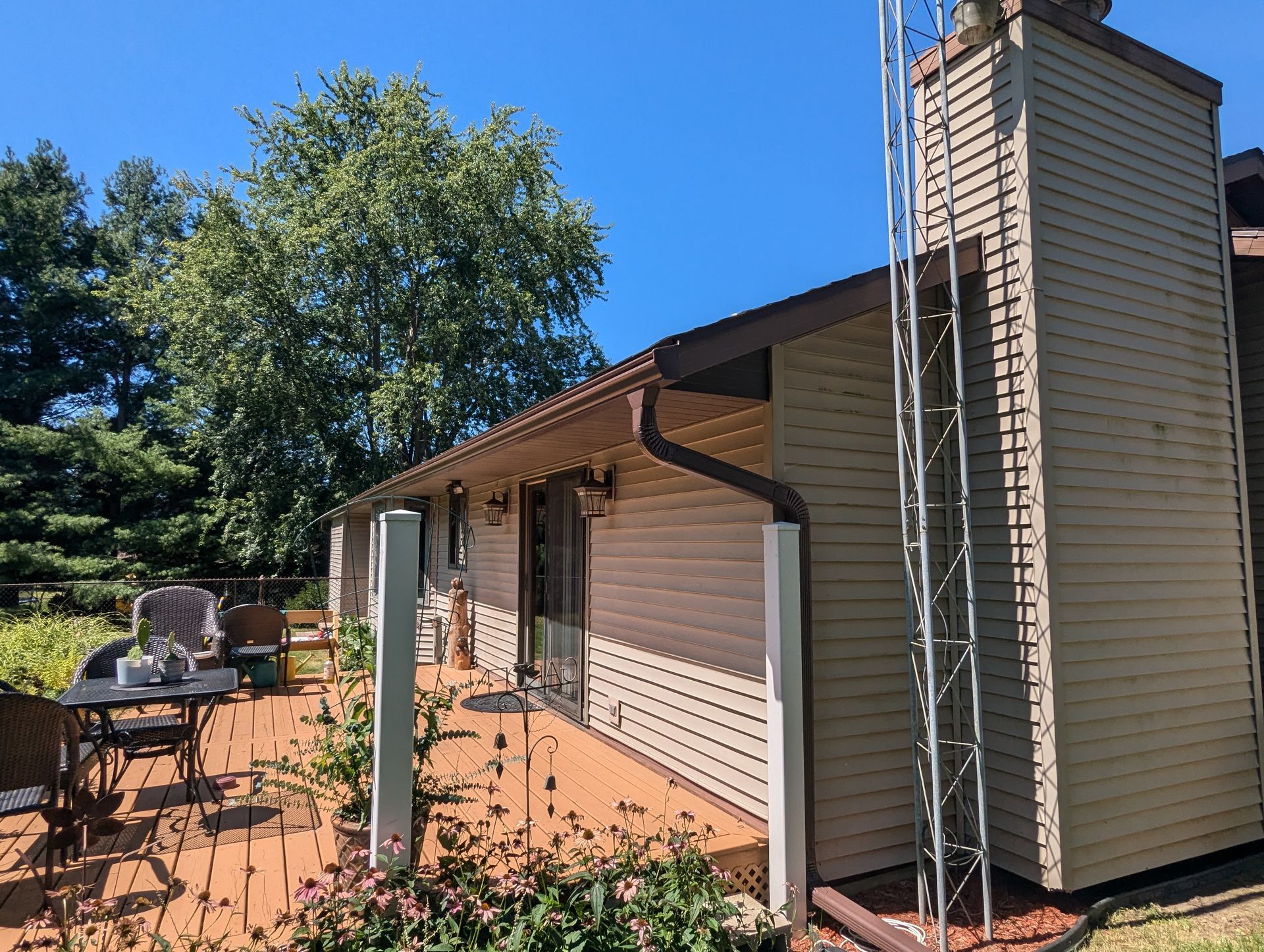 Wooden deck with table and chairs next to a light-colored building with a tall chimney and a metal pole.