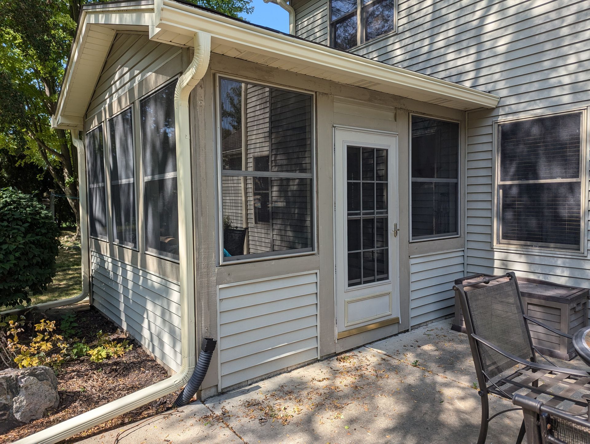 Screened-in porch attached to a house. Beige siding, windows with screens, a door. Patio furniture visible.