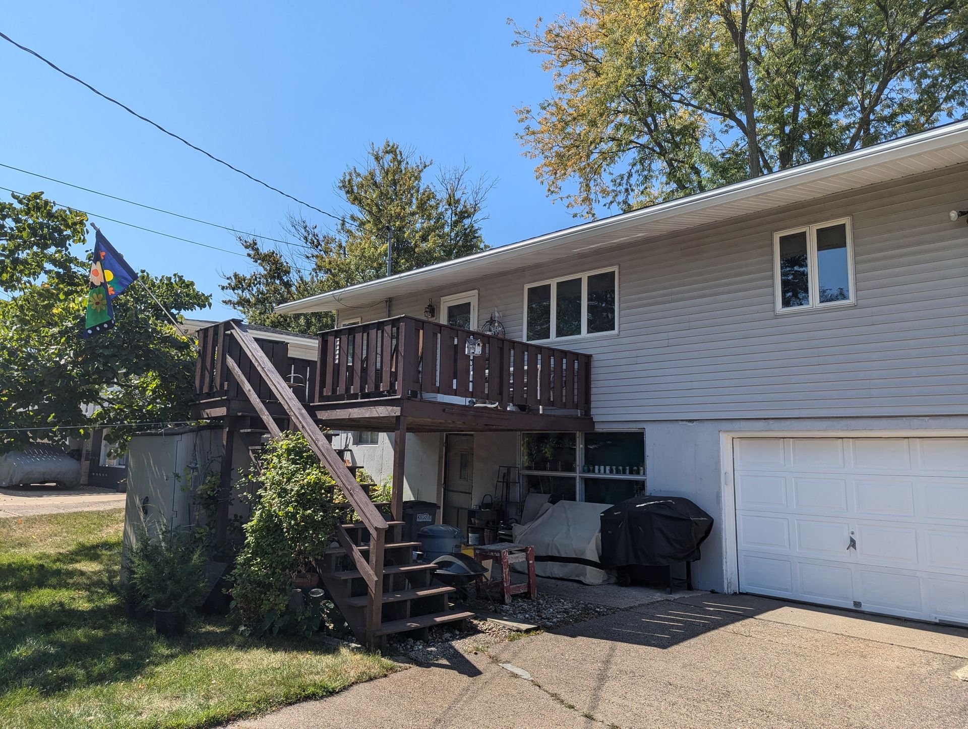 Two-story house with a wooden deck and stairs, a garage door, and a flagpole with a colorful flag.