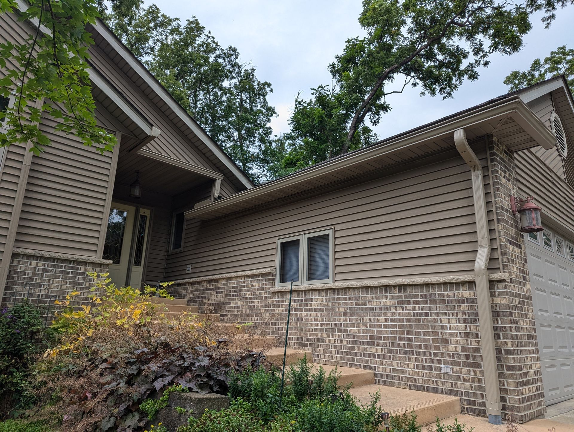 Beige house with brick, siding, and steps leading to the front door. Lush greenery and trees surround it.
