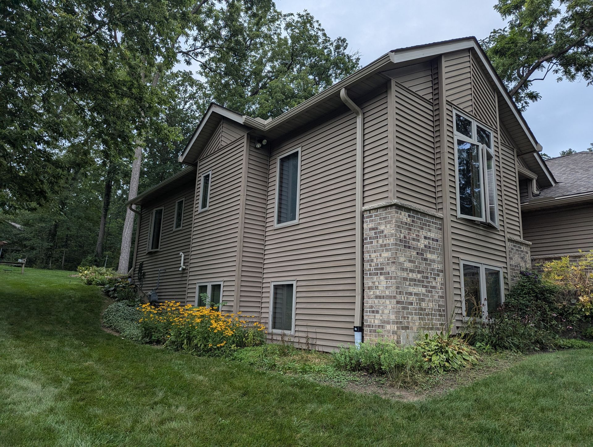 Brown house with stone accents, surrounded by greenery and trees on a grassy hill.