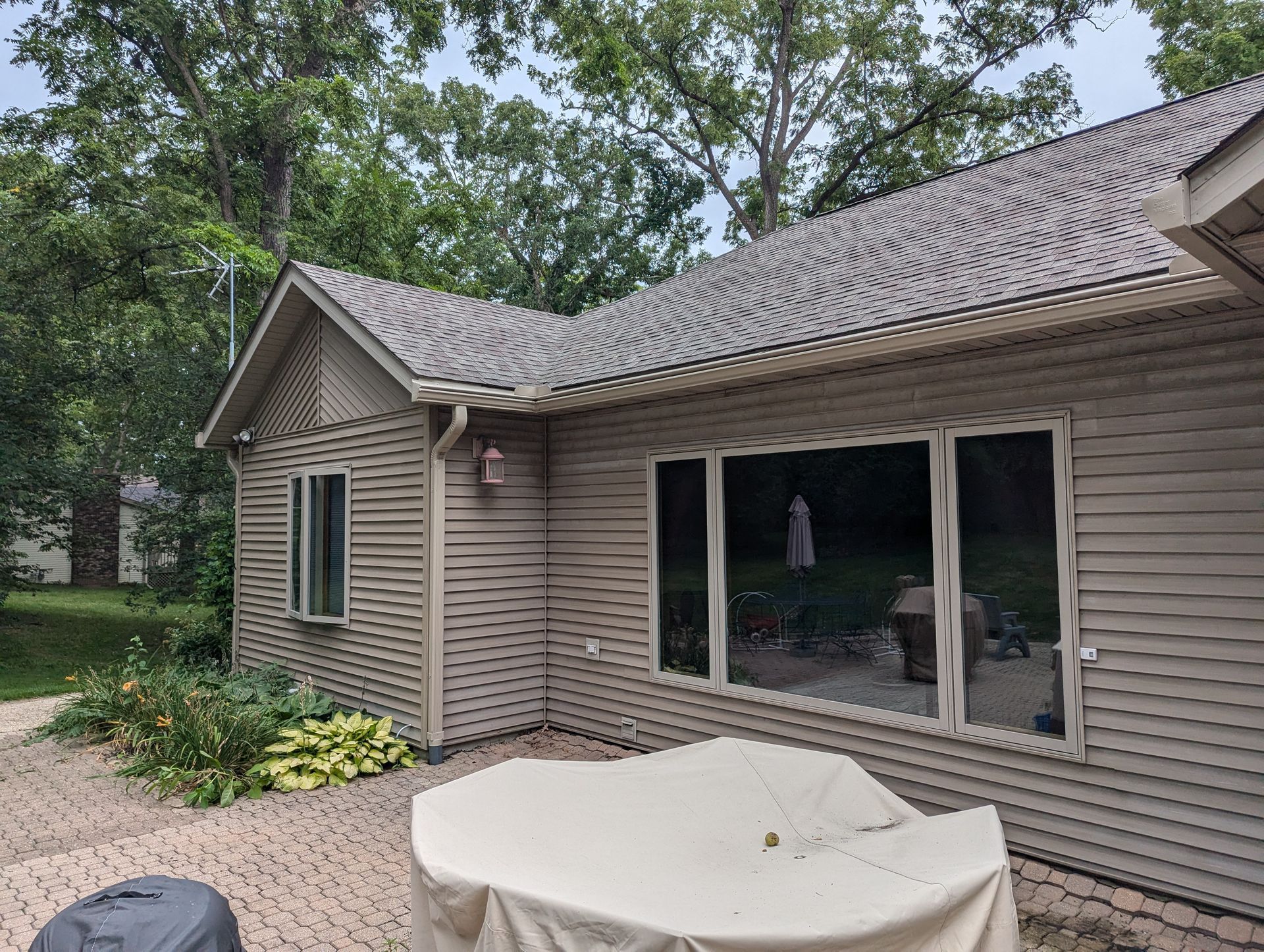 Tan-sided house with a dark roof and large windows overlooking a stone patio with covered furniture.