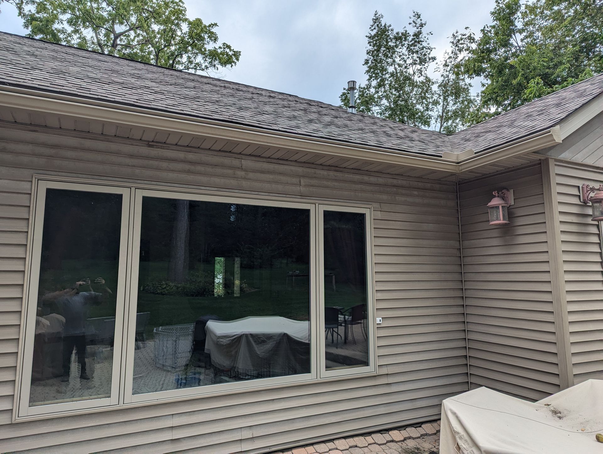 Exterior view of a house with beige siding, a large window, and a dark roof under a cloudy sky.