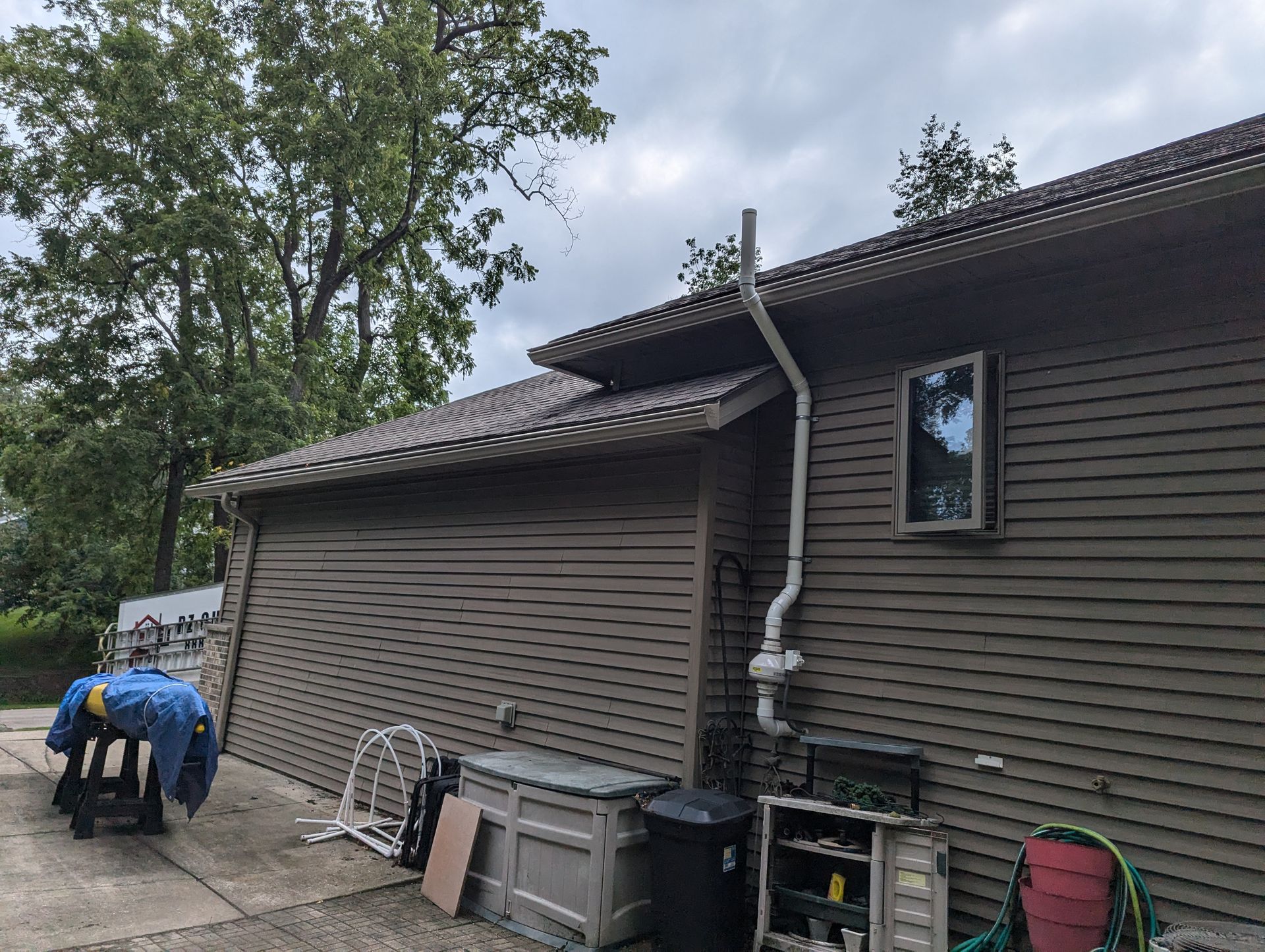 Brown siding and roof of a house with a white downspout and outdoor storage on a cloudy day.