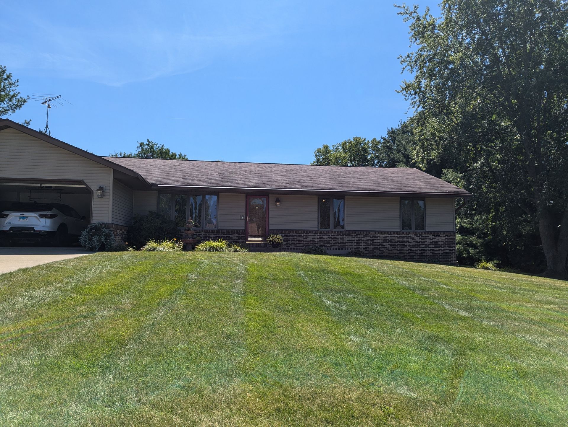 House with beige siding, brown roof, and green lawn under a blue sky. A car is in the garage.