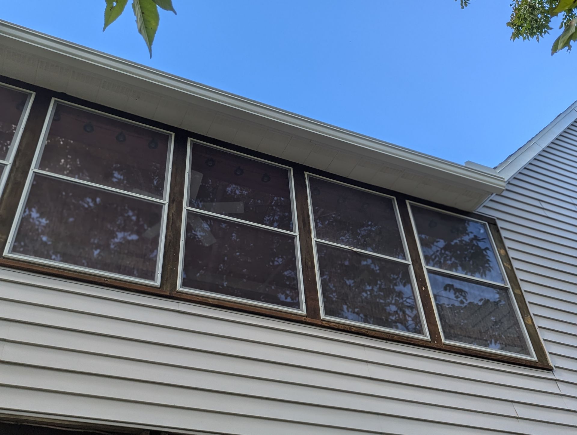 Exterior view of house with brown framed windows covered in dirty screen. White siding and blue sky.