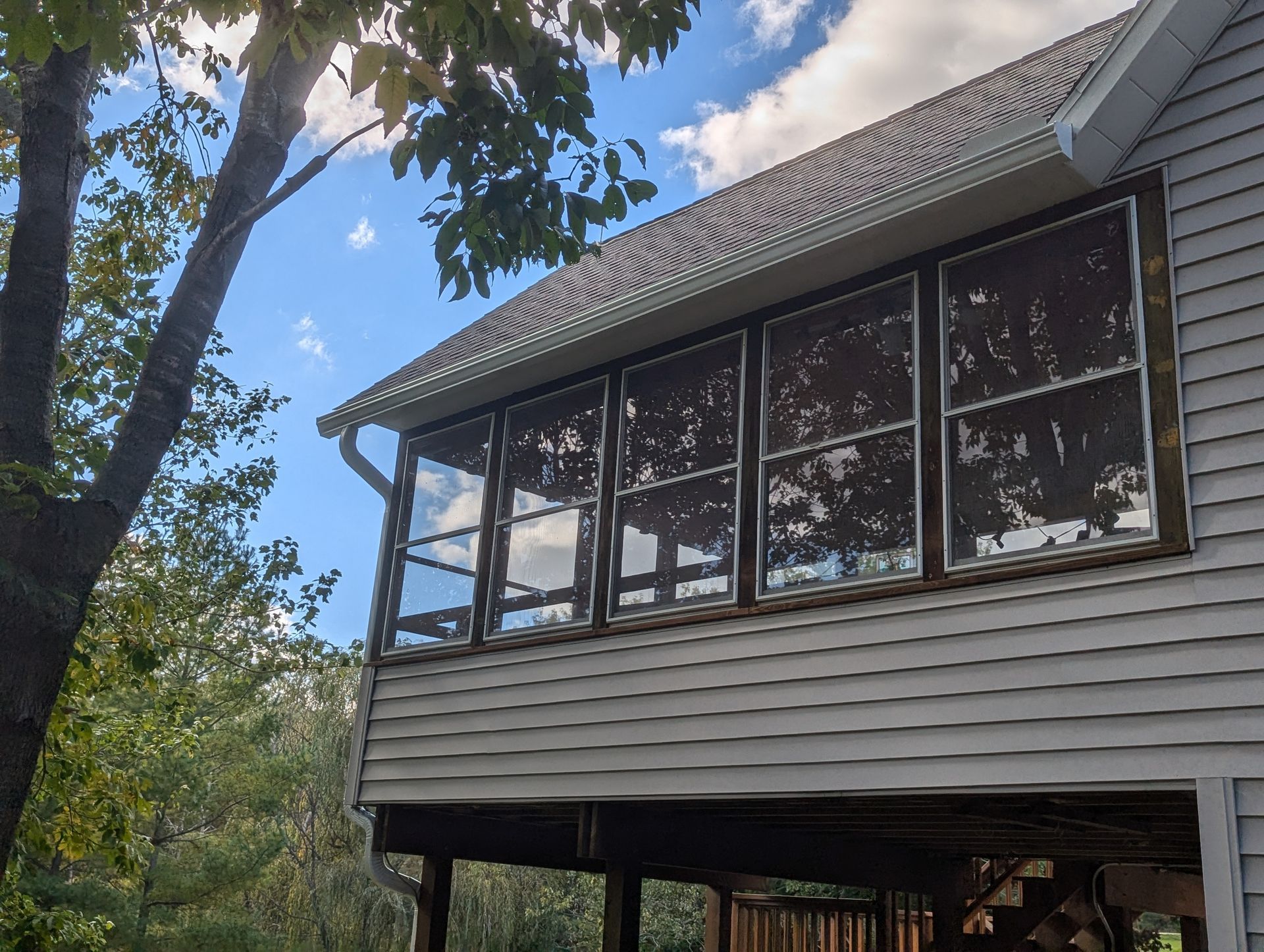 Screened-in porch on a house. Windows, white siding, gutters, a tree, and a bright sky with clouds are visible.