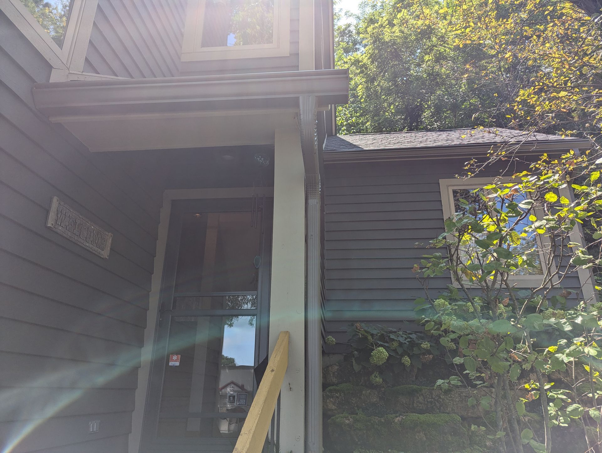 Exterior view of a gray building entrance with a door, porch, and surrounding trees in sunlight.