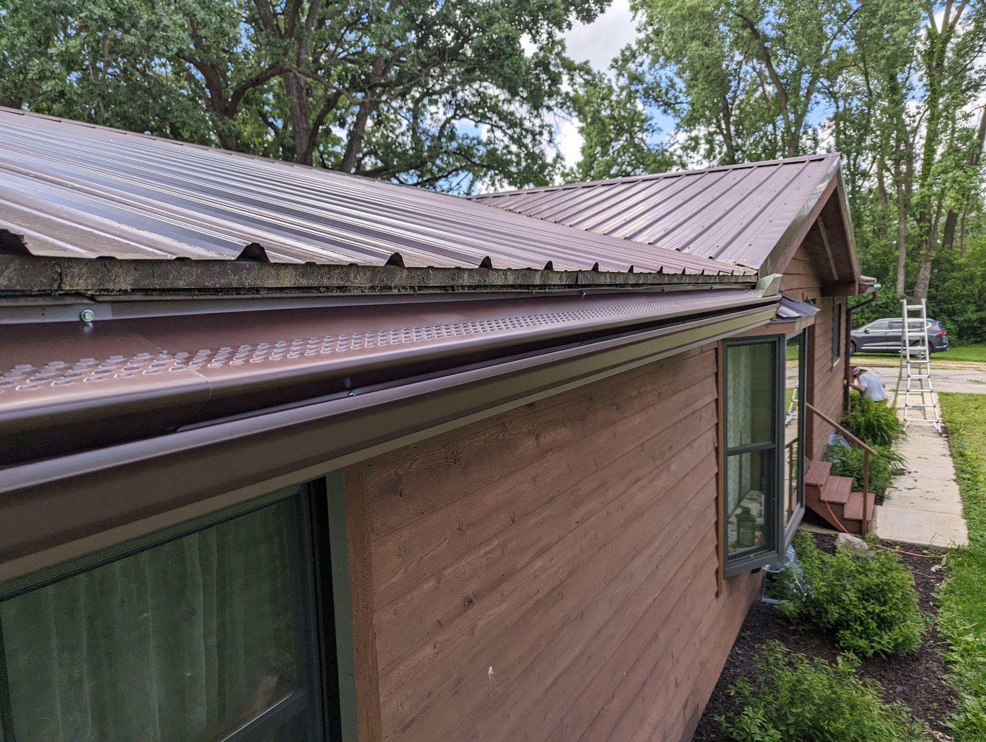 Brown metal roof and siding on a house, with gutters. Trees and sky in the background.