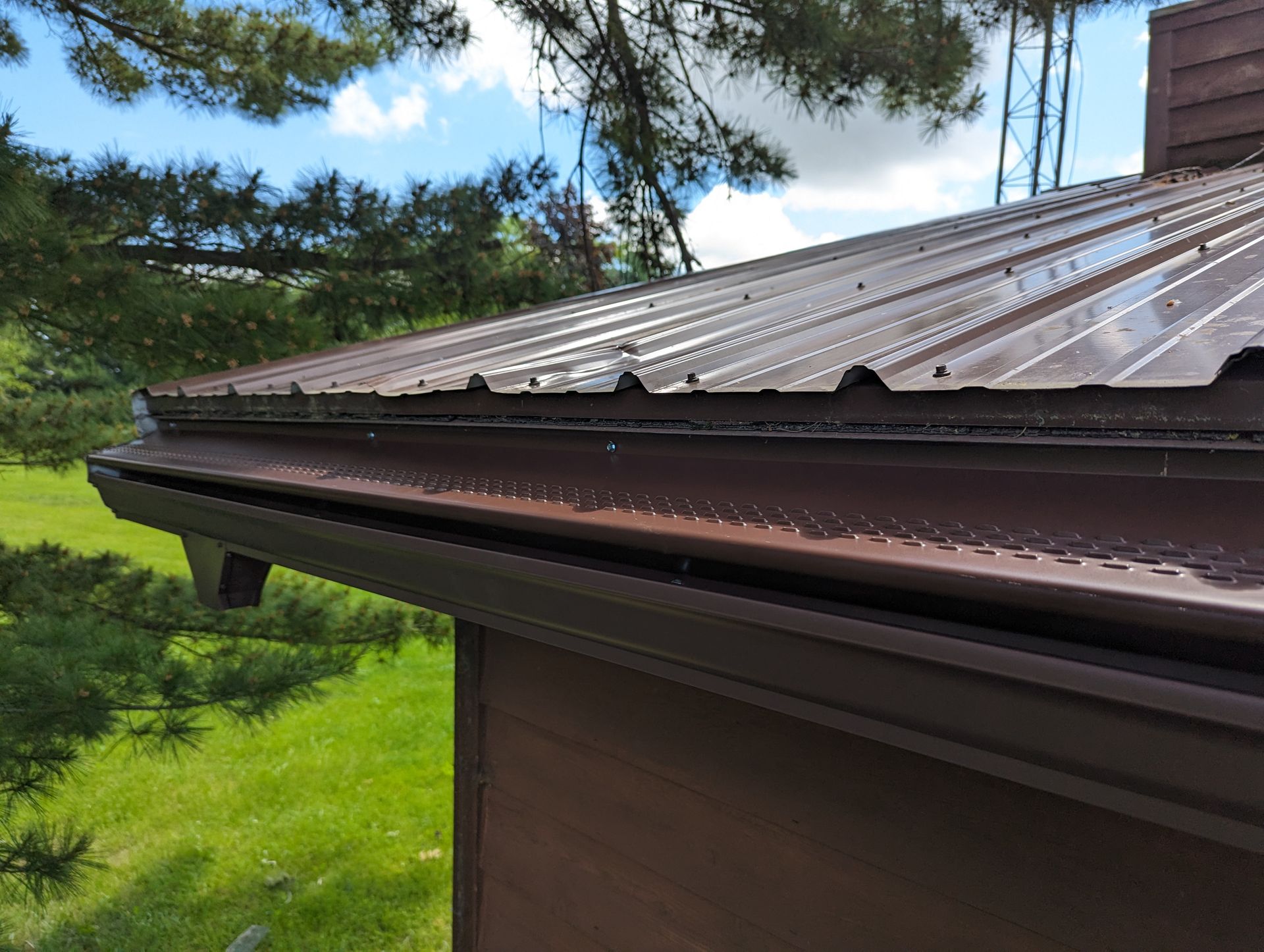 Brown metal roof and gutter, wet from rain, against a green grass and sky backdrop.