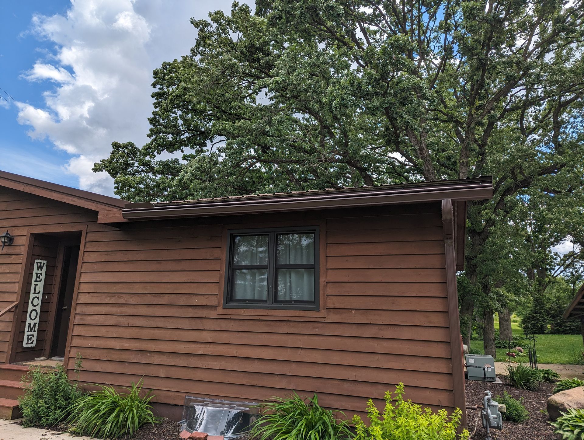 Brown house with a dark window and a tree in the background. Sunny day.
