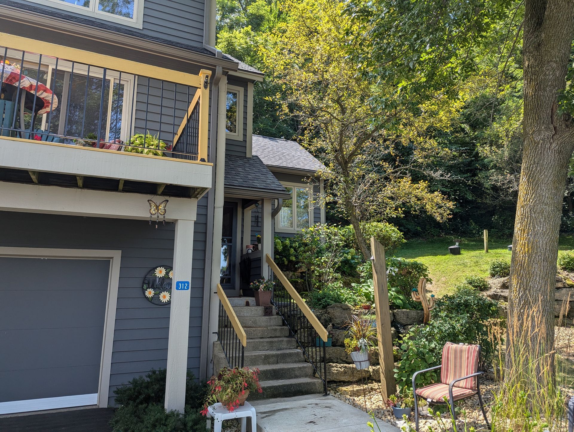 Gray house with garage, stairs to the front door, and a balcony. A tree shades a small yard.