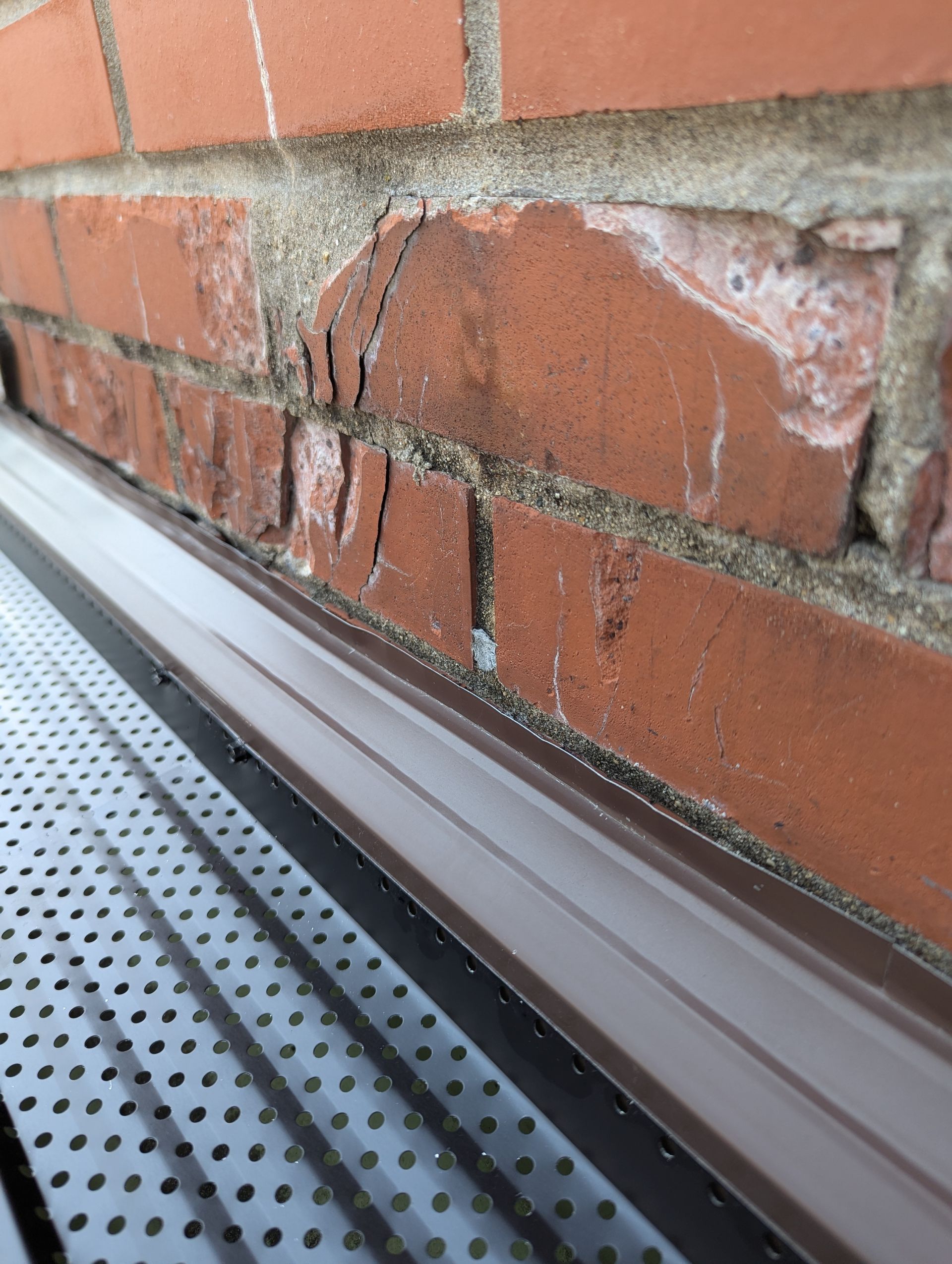 Close-up of a damaged brick wall with cracks. A gutter is below the brick.