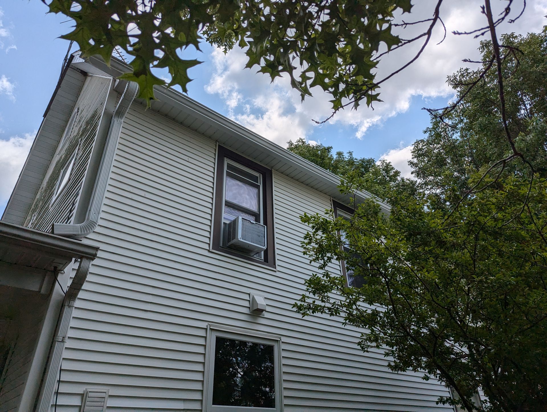 White house with air conditioner in window, under a cloudy blue sky, trees in view.