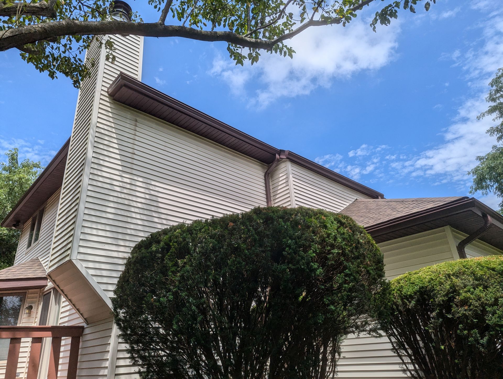 Tan-sided house with brown roof and chimney against a blue sky, topped by a tree branch and shrubs.