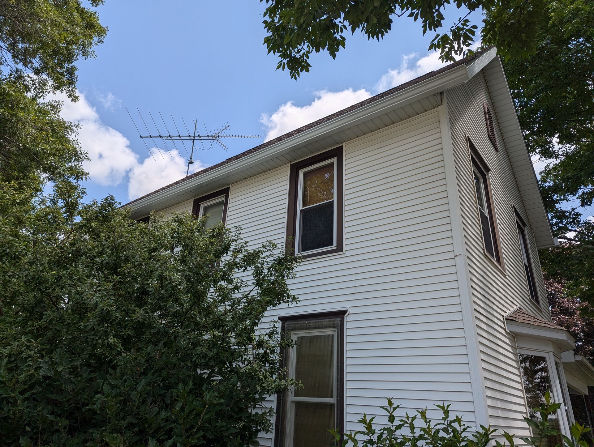 White house with dark window frames and TV antenna against a blue sky, framed by trees.
