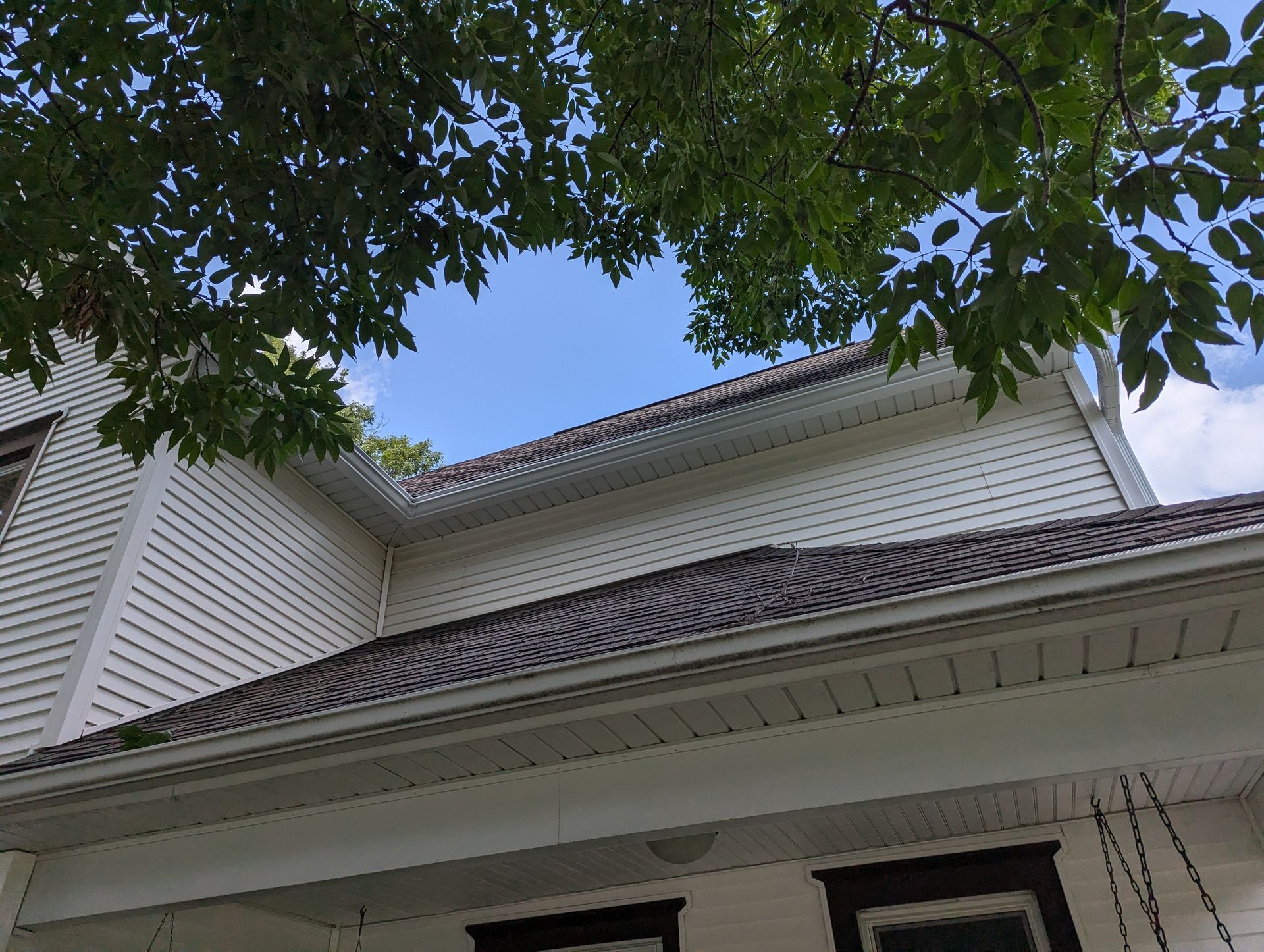 White house exterior with dark roof under a blue sky, viewed from below with leafy tree branches overhead.