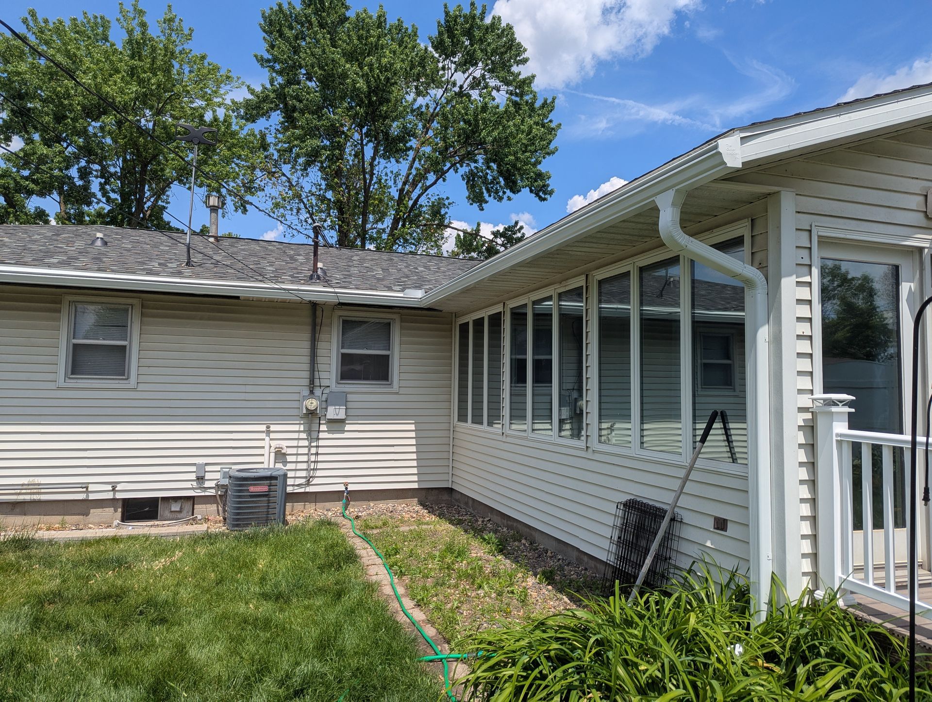 Beige house with a sunroom, gutters, and a grassy yard under a blue sky.