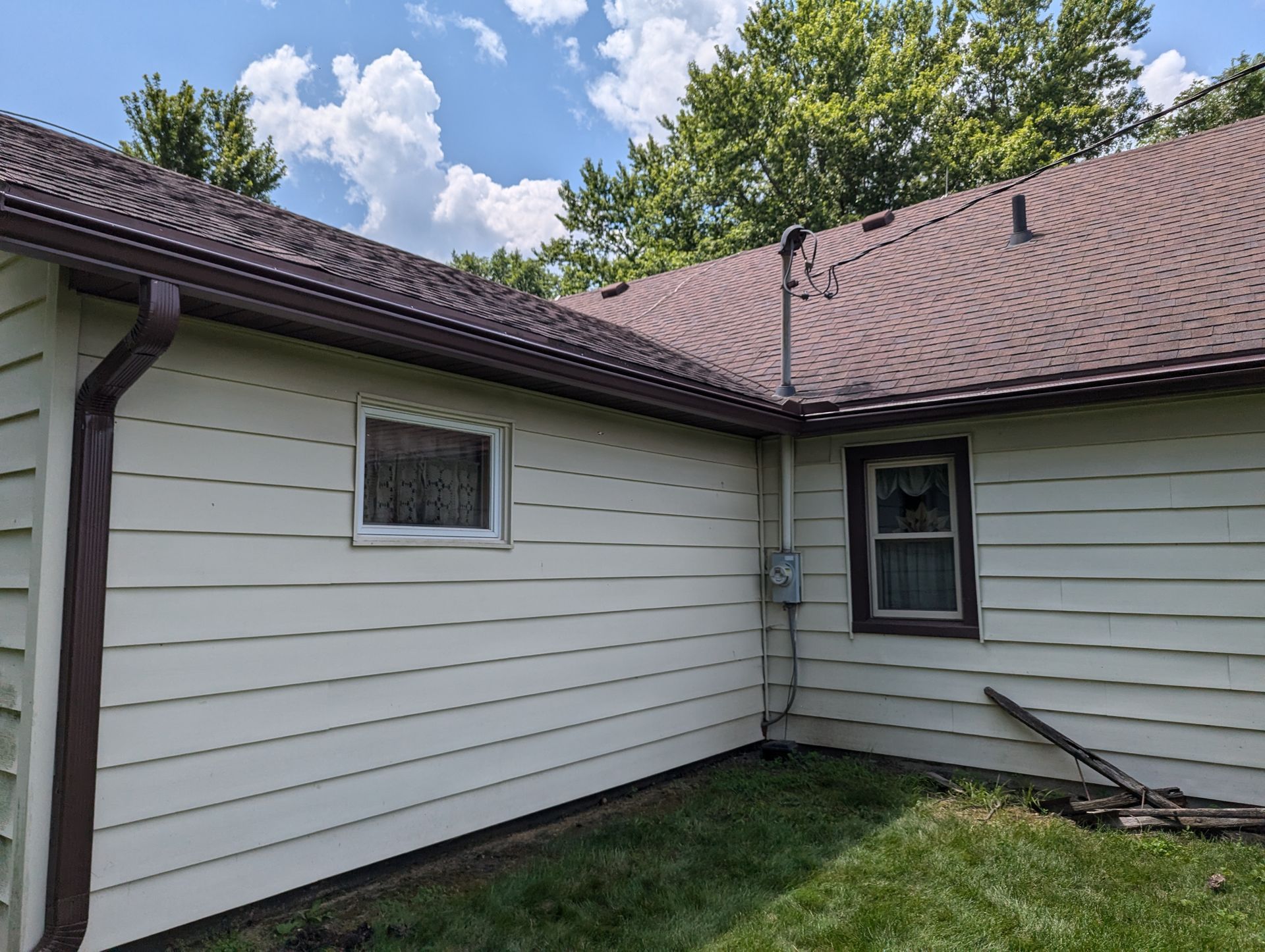 Side view of a house with light green siding and a brown roof, gutters, and window trim, set against a cloudy blue sky.
