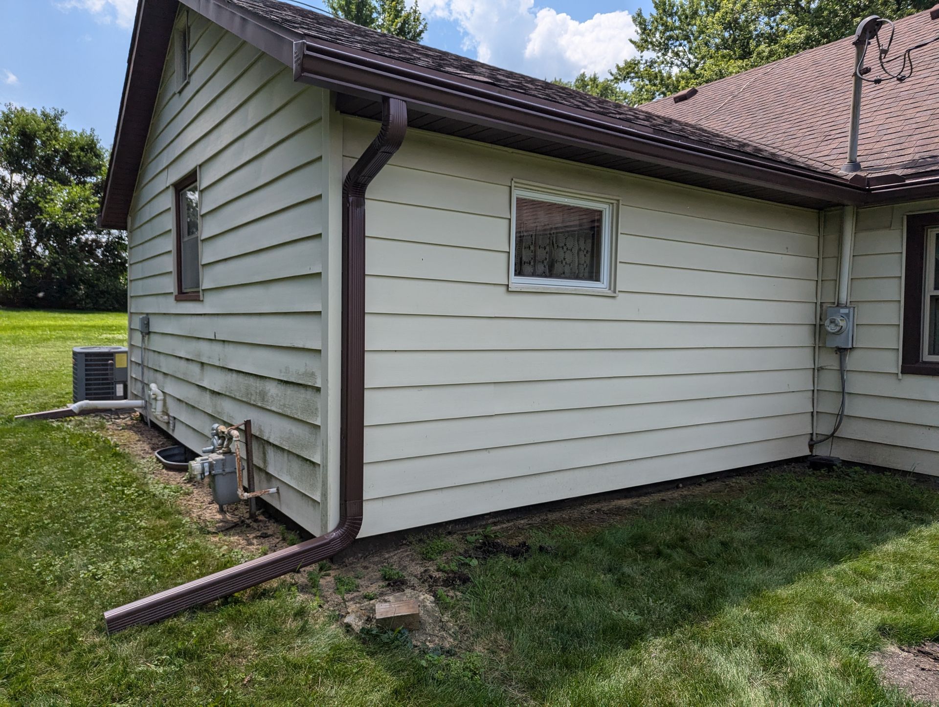 Exterior of a house with light green siding, brown gutters, and windows on a grassy lawn.