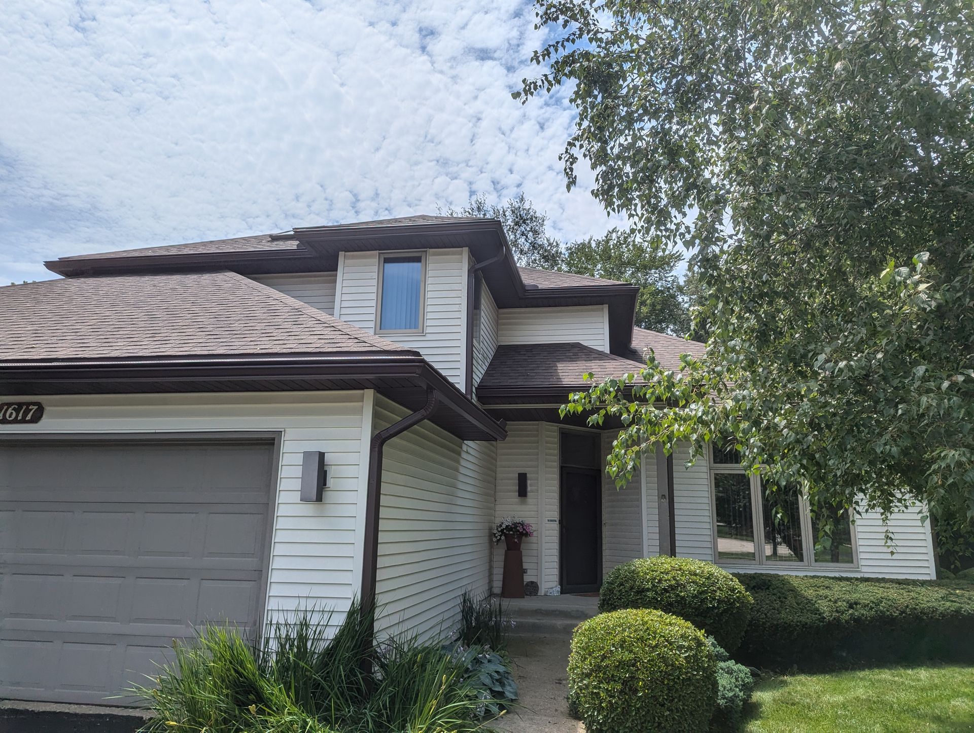 Two-story white house with brown roof and garage, front door under a small porch, bushes and a tree.
