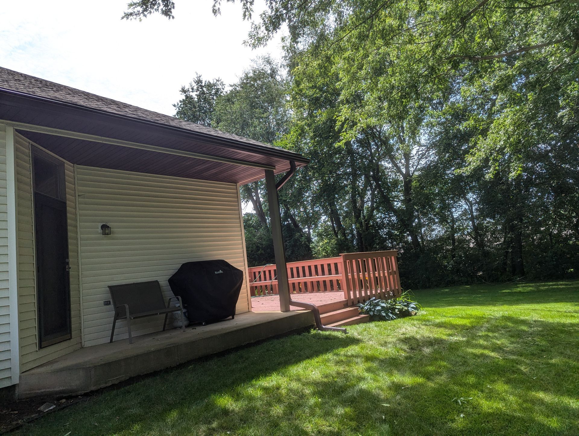Backyard with deck and grill next to house with light siding and brown trim, surrounded by green grass and trees.