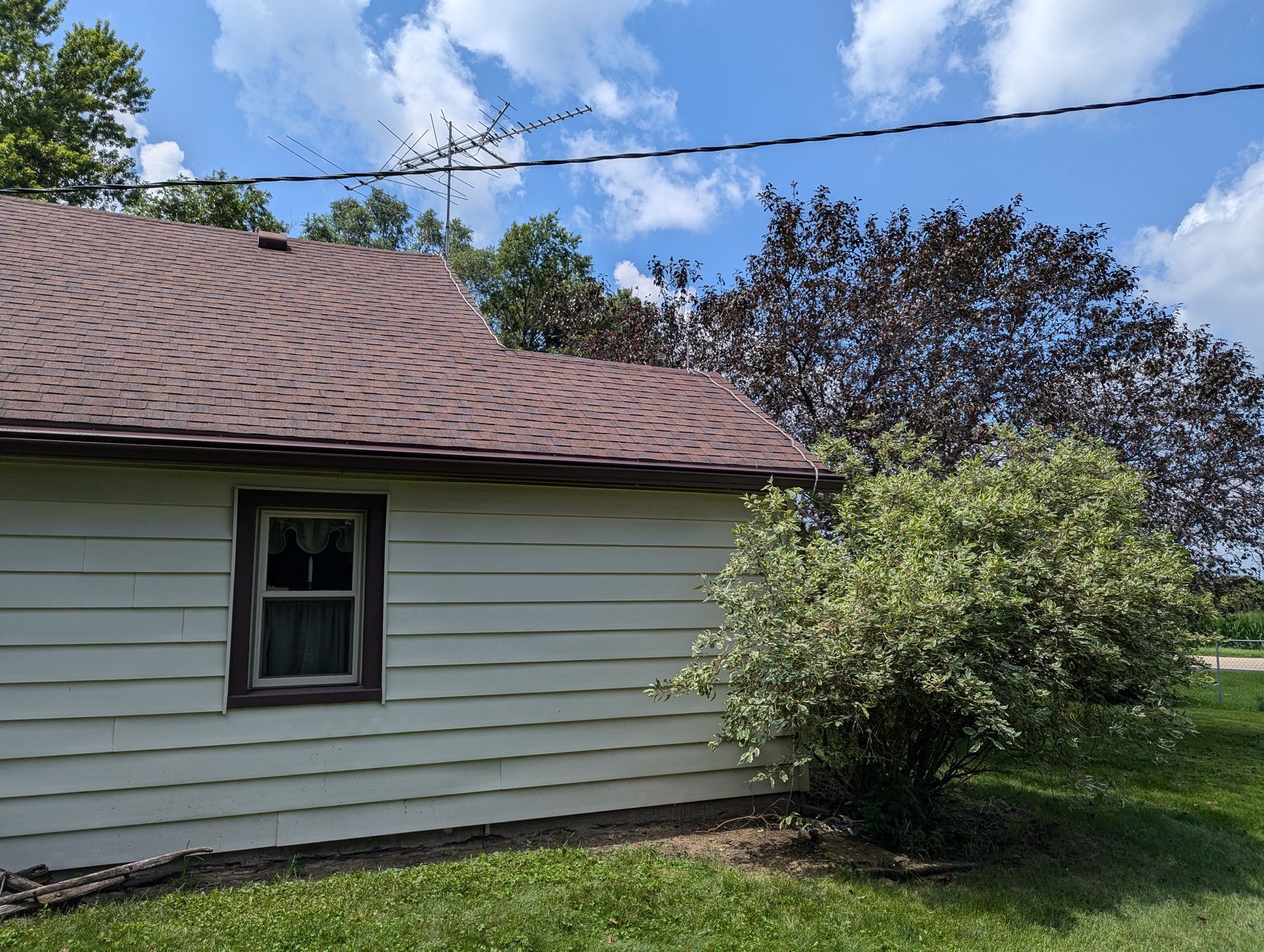 A white house with a brown roof and a small window next to a green bush and a tree with purple leaves under a blue sky.