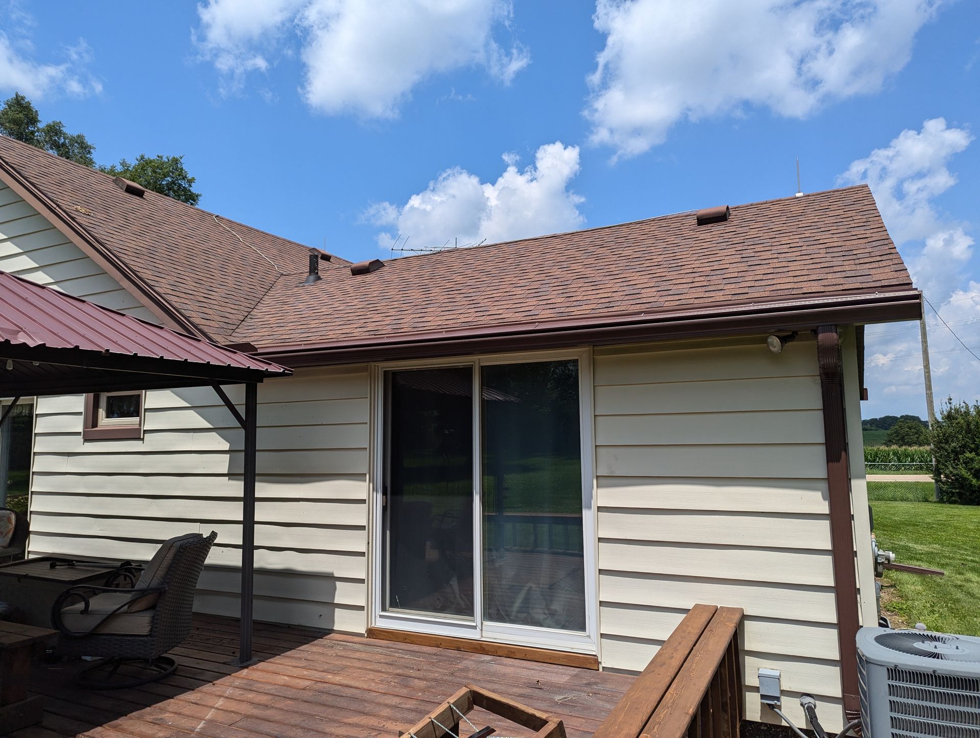 Beige house with brown roof, sliding glass door, and wooden deck against a blue sky with clouds.